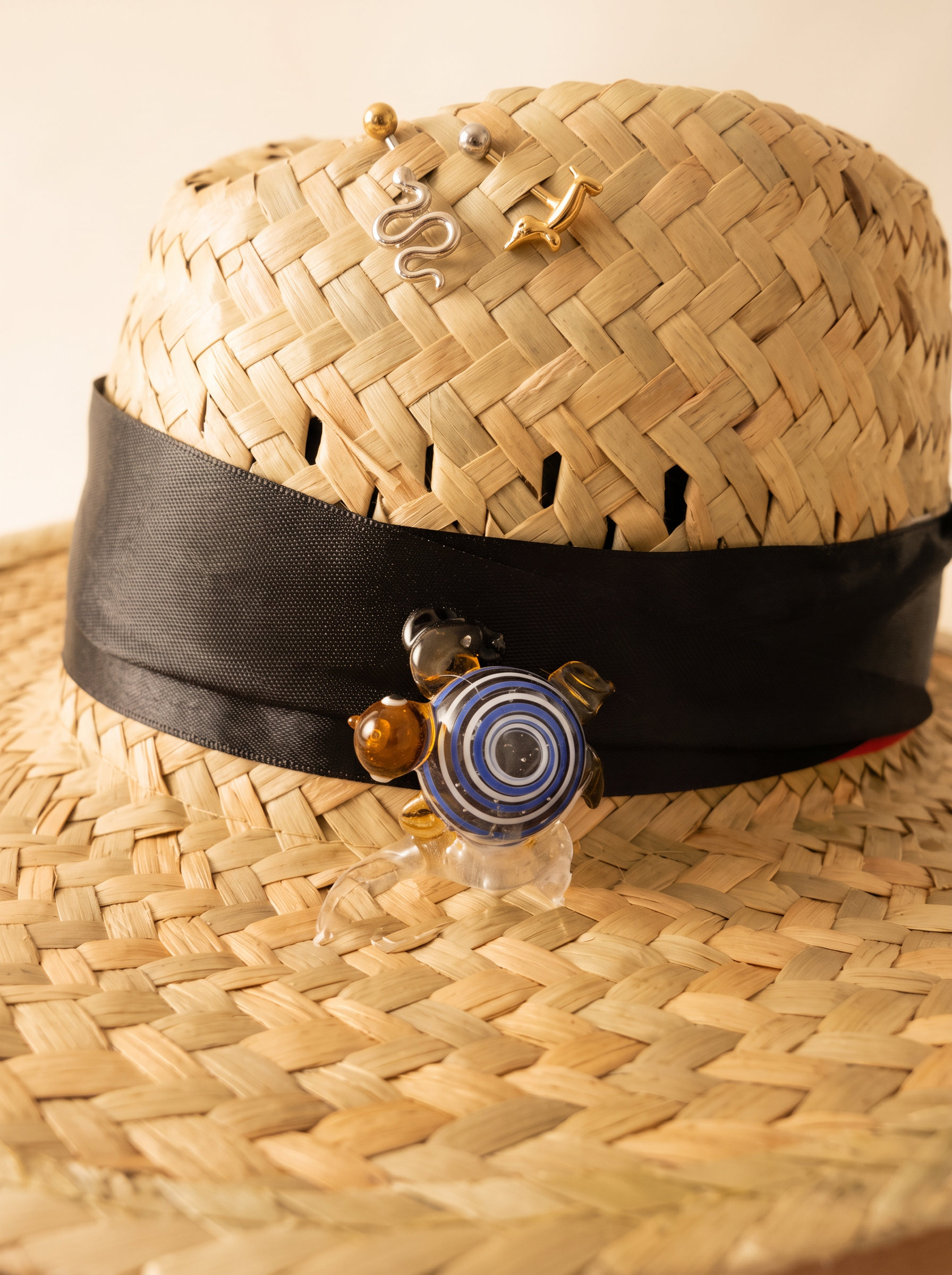 Macro detail of a straw hat with black band, silver snake and gold dolphin pins and a blue glass turtle charm
