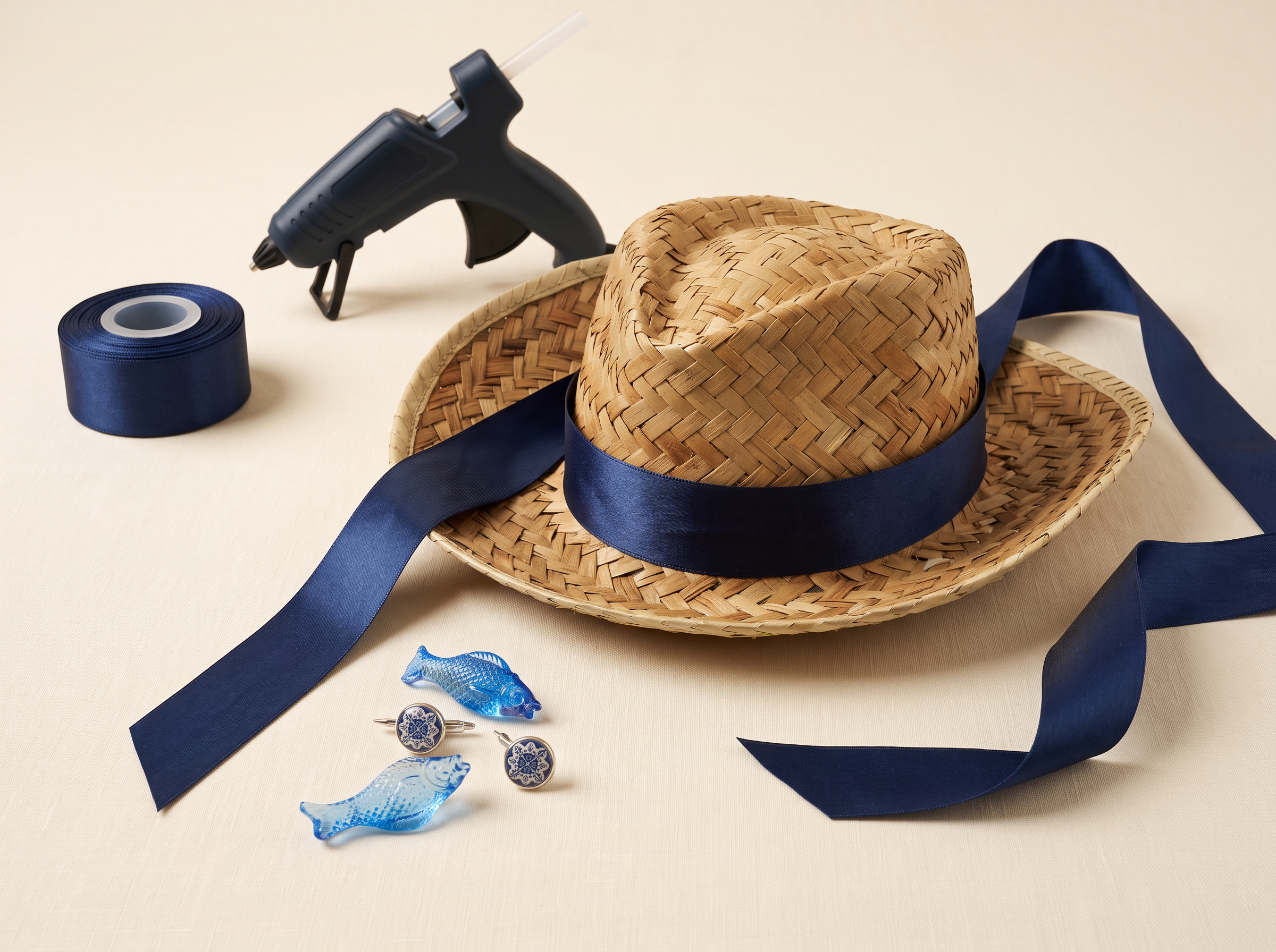Straw hat, navy satin ribbon, glue gun and blue fish and azulejo charms laid out on a cream studio surface