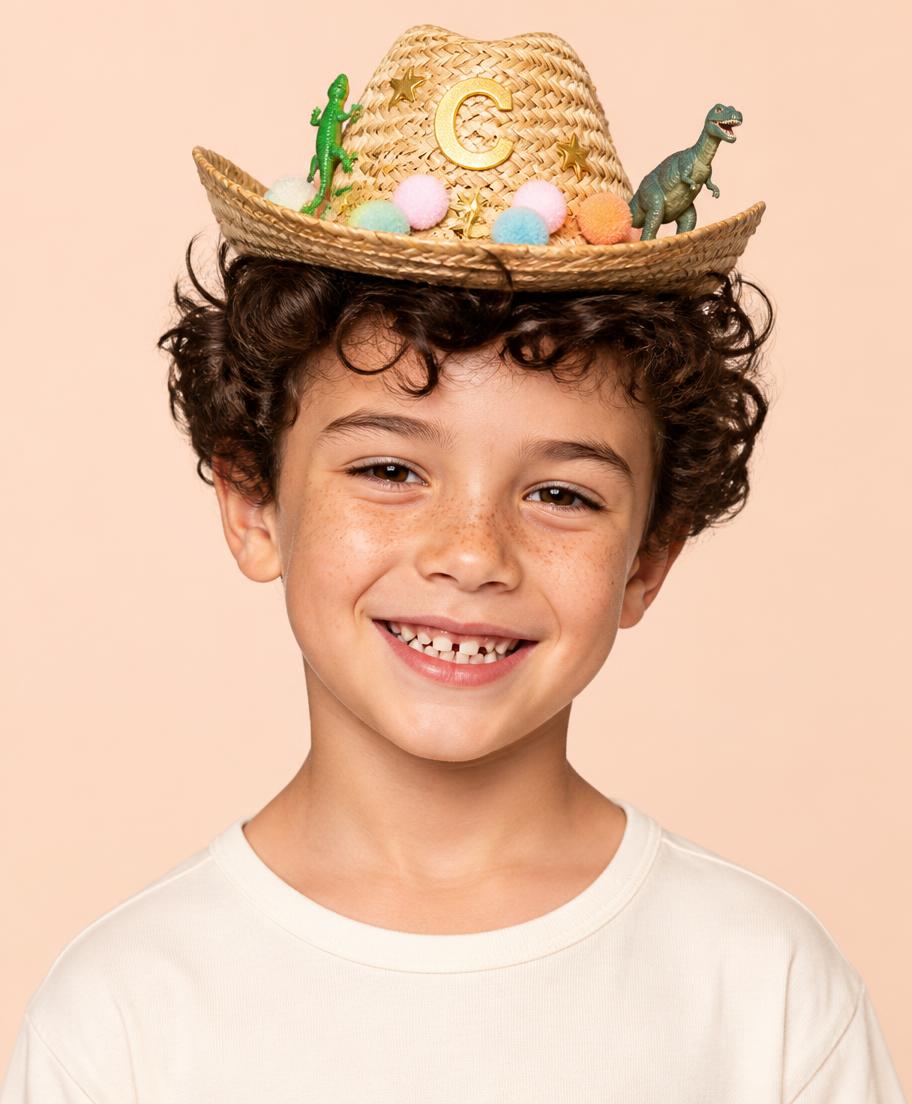 Six year old boy with curly hair grinning in a mini straw cowboy hat with a gecko, dinosaur, pastel pom poms and a gold letter C
